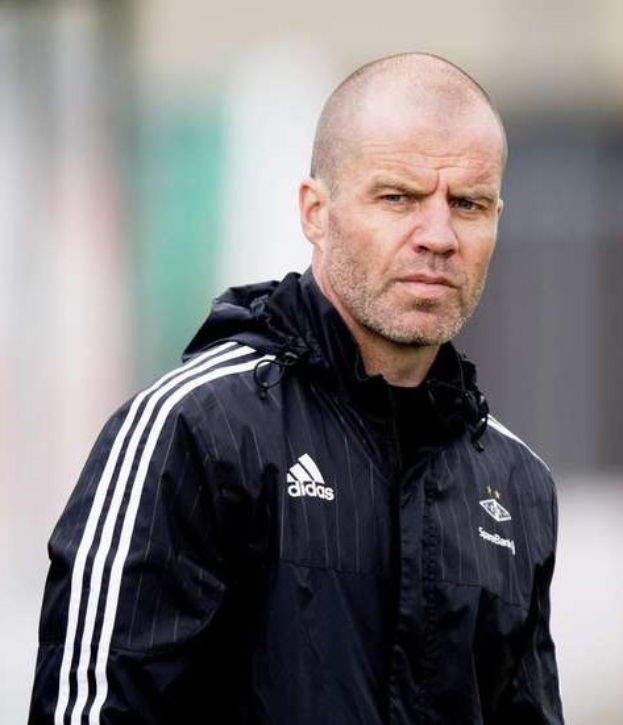 Baldiger Fußballtrainer in marineblauer Jacke mit weißen Streifen und weißem Kragen, der auf einem Fußballfeld steht und nachdenklich wirkt.