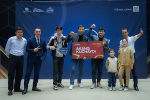 Gruppe von Menschen auf einer Bühne, die eine Tafel mit der Aufschrift "Akang Kuchaydi - Zakovat Quiz" halten, mit einem Banner, Barcode und Lichtern im Hintergrund.