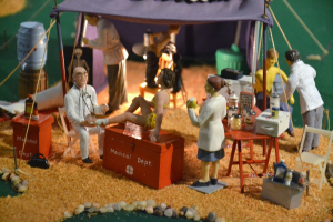 Eine Puppe steht auf einer Box mit der Aufschrift 'medizinisches Departement' mit einem Tisch voller Gegenstände in der Nähe, alles unter einem violetten Zelt, mit zusätzlichen Gegenständen im Hintergrund.