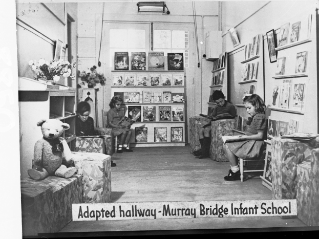 Eine Gruppe von Kindern sitzt auf Stühlen in einem Raum mit Bücherregalen, einem Teddybär, einer Blumenvase auf einem Tisch und gerahmten Gegenständen an der Wand, mit dem Text "Angepasster Flur Murray Bridge Kindergarten" unten.