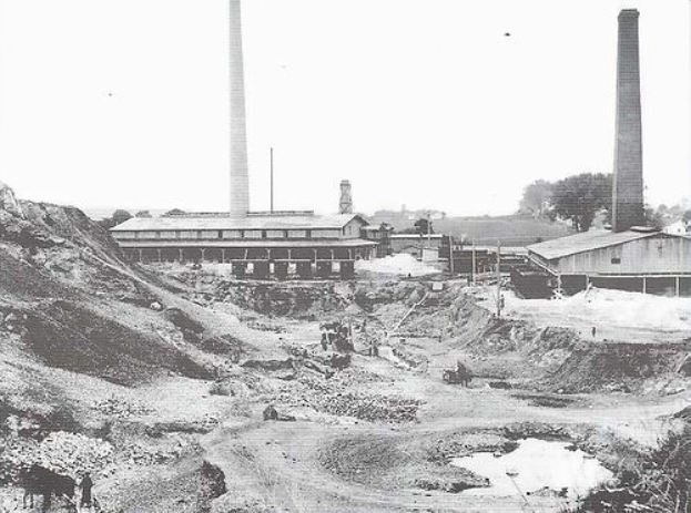 Ein Schwarz-Weiß-Foto einer Baustelle mit zwei Schornsteinen im Hintergrund, umgeben von Gebäuden, Masten, Bäumen und einem klaren Himmel, mit Text am unteren Rand, der darauf hinweist, dass es sich um ein kohlebetriebenes Kraftwerk handelt.