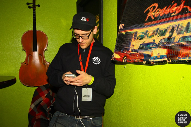 Eine Person mit einer Mütze und einem Ausweis hält ein Mobiltelefon in der Hand, neben ihr eine Gitarre und rechts daneben ein Plakat.