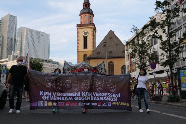 Eine Gruppe von maskierten Menschen mit einer Fahne, die durch eine Straße marschieren, mit einem geparkten Auto auf der linken Seite, Gebäuden und einem Uhrenturm im Hintergrund bei klarem blauem Himmel.