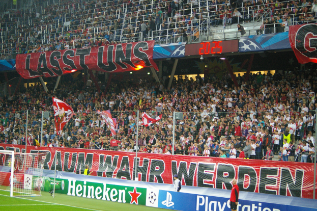 Ein Fußballspiel in einem Stadion mit einer großen Menge, grünem Rasen, einem Torpfosten links, Bodenbannern, Menschen mit Fahnen und Hintergrundgeländern, Tafeln, Lampen und anderen Gegenständen.