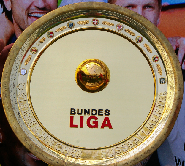 Ein Mann hält einen goldenen Schild mit 'Bundesliga' darauf, im Hintergrund eine Fahne mit drei Personen