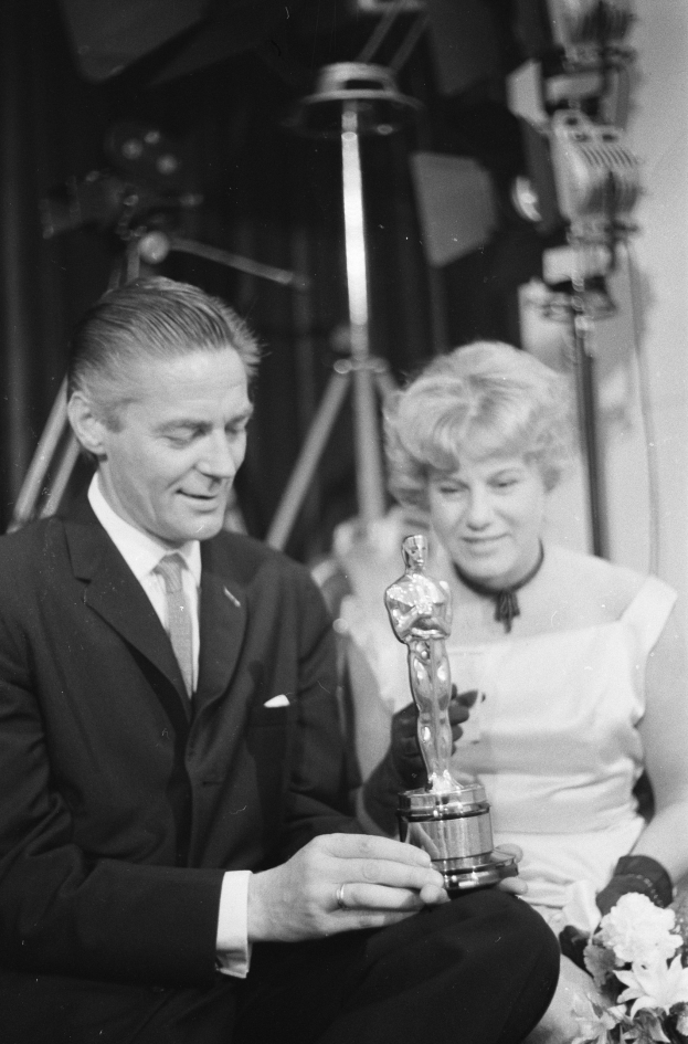 Ein Mann in einem schwarzen Blazer, weißem Hemd und Krawatte hält eine Oscar-Statue, waß er neben einer Frau in Abendgarderobe sitzt, die einen Blumenstrauß hält, mit Hintergrundbeleuchtung, die die schwarz-weiße Szene erhellt.