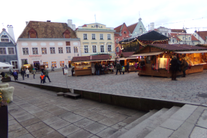 Ein lebendiger Weihnachtsmarkt in Tallinn, Estland, mit Menschen um beleuchtete und geschmückte Stände, Gebäuden mit Fenstern im Hintergrund, Treppen, die zum Markt führen, und Töpfen mit Pflanzen.