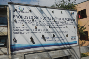 Reklame an der Seite eines Gebäudes mit der Aufschrift "Vorgeschlagen 2016 Total Pipe Replacement" im Vordergrund, mit Gebäuden, Bäumen, Drähten und einem klaren blauen Himmel im Hintergrund.
