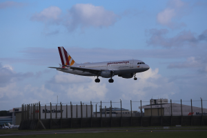 Germanwings Airbus A320-200 startet von Frankfurt Airport mit Gras darunter, einem Zaun im Vordergrund, Gebäuden und Bäumen im Hintergrund und einer bewölkten Himmel.