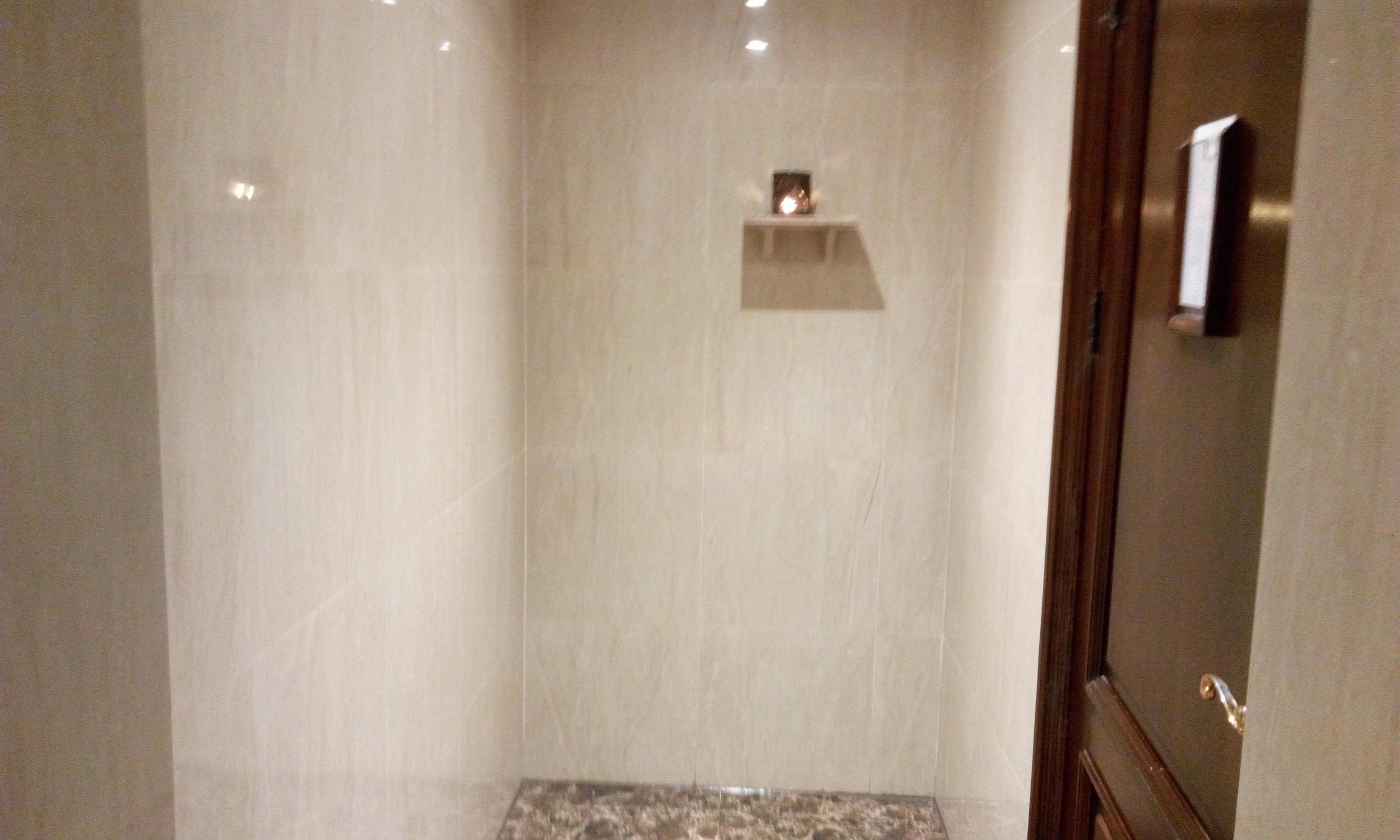 Ein Badezimmer mit einer Walk-in-Dusche, einer Toilette und gefliesten Wänden, das eine Deckenbeleuchtung und eine Tür auf der rechten Seite aufweist.