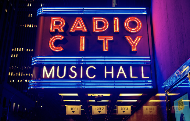 Radio City Music Hall in New York City, hell erleuchtet in der Nacht, umgeben von hohen Gebäuden und einem 'Radio City'-Schild auf der rechten Seite mit zusätzlichen Tafeln darunter.