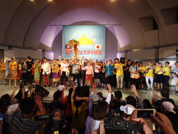 Eine Gruppe von Menschen auf einer Bühne mit Kameras, Handys und Musikinstrumenten während einer Eröffnungszeremonie eines Festivals, mit einem Banner, Wänden und Deckenlampen im Hintergrund.