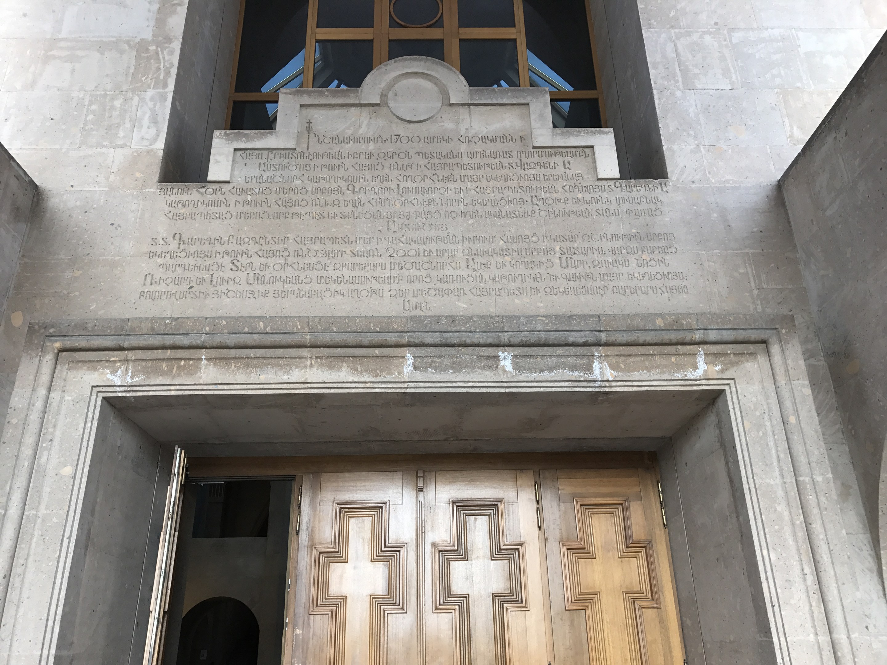 Große hölzerne Tür vor einem Steinbau mit einem Glasfenster oben und einer Inschrift an der Wand, wahrscheinlich ein Holocaust-Mahnmal.