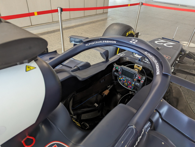 Cockpit eines Formel-E-Autos in einer Ausstellung mit Lenkrad im Vordergrund, Wand im Hintergrund, Auto auf dem Boden und gespannten Pfosten.