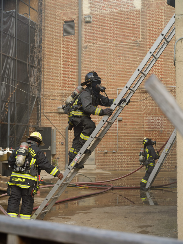 Feuerwehrleute mit Helmen und Luftzylindern klettern an einer Leiter vor einem Backsteingebäude mit Rohren auf dem Boden und einem Metallstab darunter.