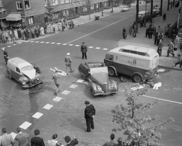 Schwarze und Weiße Szenerie eines Autounfalls an einer Straßenecke mit mehreren beteiligten Fahrzeugen und einer Gruppe von Menschen in der Nähe, mit Gebäuden, Bäumen und Schildern im Hintergrund.