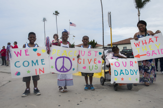 Eine Gruppe von Kindern hält Schilder in der Hand und steht vor einem Gebäude, während eine Frau einen Kinderwagen schiebt, in dem ein Baby sitzt; auf einem Schild steht 'Wir kommen in Frieden, gebt uns unseren Daddy.'