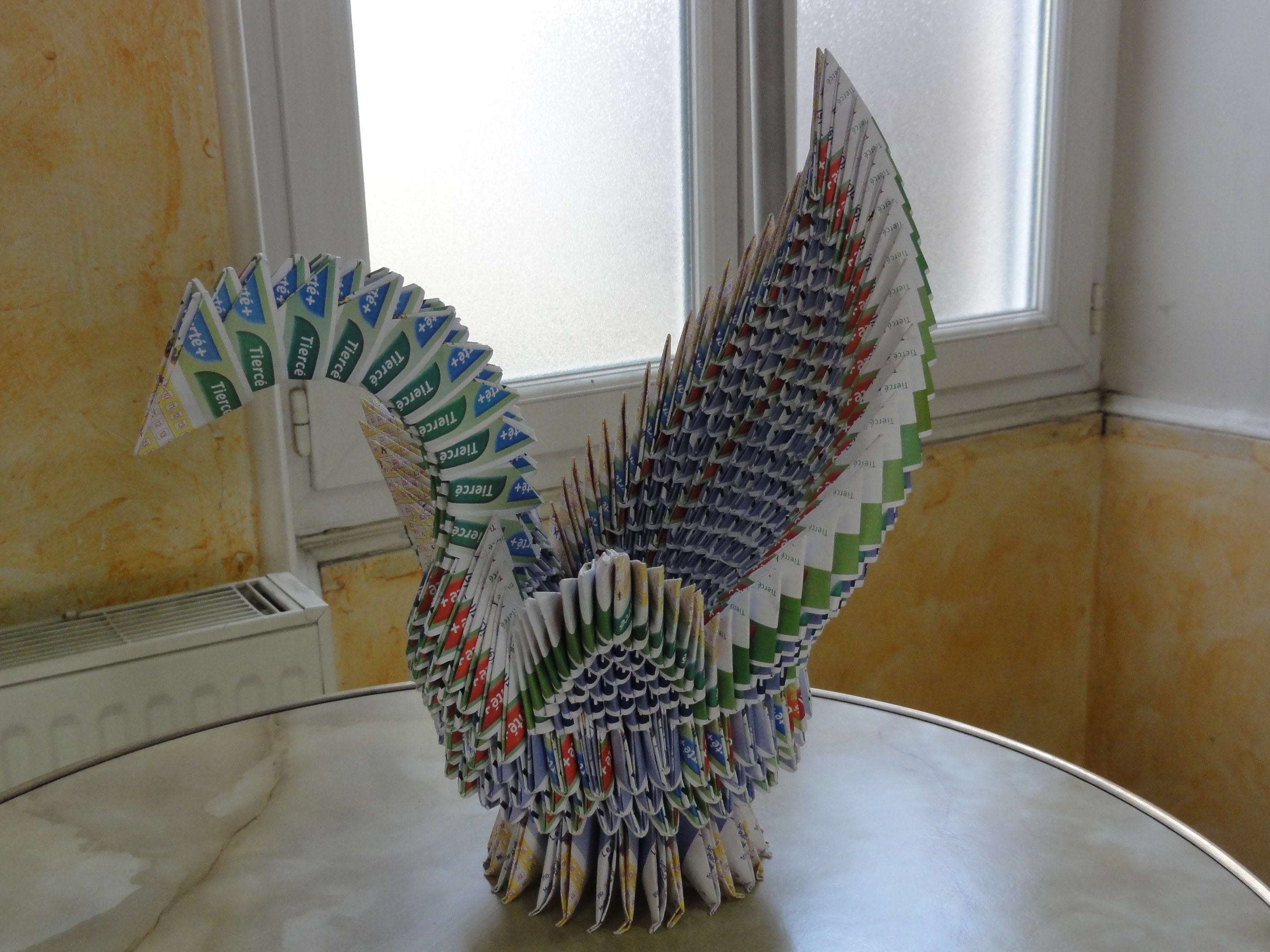Komplexe Origami-Pfau aus Toilettenpapier, auf einem Tisch gegen eine Wand mit einem Fenster gestellt.