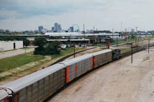 Güterzug fährt auf Schienen neben einer Stadtlandschaft mit Strommasten, Kabeln, Gebäuden, Bäumen, Gras und bewölktem Himmel.