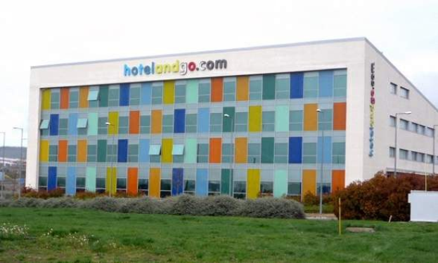 Großes modernes Hotelgebäude mit vielen Fenstern, umgeben von Grünflächen, Fahrzeugen und einem bewölkten Himmel, mit "hotel und go"-Schriftzug.