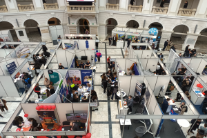 Eine große Gruppe von Menschen versammelt sich in einem Raum voller Stände, Banner, Tische, Stühle und Ballons, mit einem Gebäude im Hintergrund, das Bögen, Säulen, Geländer und einen Bildschirm aufweist.