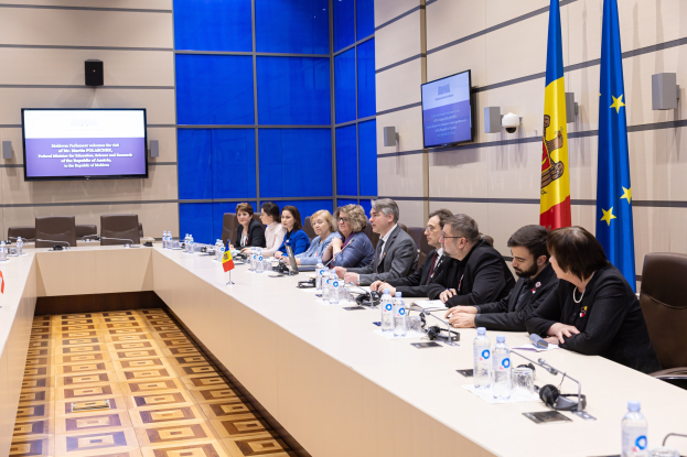 Gruppe von Menschen, die um einen langen Tisch in einem Konferenzraum sitzen, mit Flaschen, Gl├Ąsern, Mikrofonen, Papieren und Fahnen im Hintergrund, mit Fernsehern und Lautsprechern an der Wand dahinter.