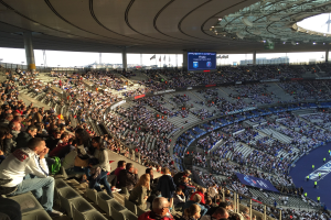 Große Zuschauermenge in einem Stadion bei einem Fußballspiel, mit einer Bühne auf der rechten Seite, Fahnen, Stangen, einem Bildschirm und der Allianz Arena in München, Deutschland im Hintergrund.